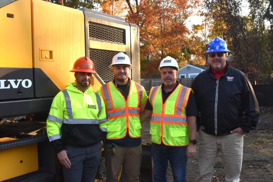 Burns Construction crew of four smiling employees in hard hats standing in front of a Burns excavator